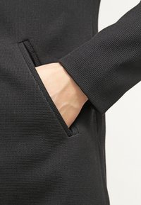 Musta kuvioitu takki, jossa on tasku ja käsi lepää sisällä, korostaen kankaan kudottua kuviota ja jäsenneltyä muotoilua.