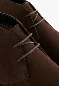 Des chaussures en daim marron avec deux œillets pour les lacets et des lacets assortis. La texture est douce avec des coutures détaillées le long des bords.