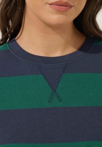 Lähikuva henkilön alaleuasta ja kaulasta, joka on pukeutunut laivastonsiniseksi ja vihreäksi raidalliseen pyöreäkaulaiseen villapaitaan, jossa on ommeltu V-yksityiskohta kauluksessa.