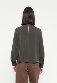Blouse vert olive à manches longues, coupe décontractée, avec détail œillet au dos. Le tissu semble lisse ; associée à un pantalon marron, créant un look décontracté.