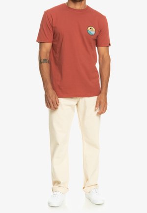 T-shirt en coton rouge à manches courtes, col rond, et logo circulaire représentant des vagues. Associée à un pantalon clair et des baskets blanches.