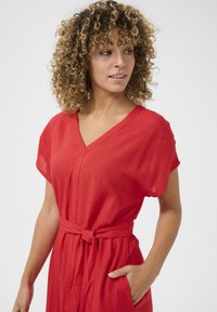 Röd klänning med kort ärm och V-ringning, med bälte i midjan och sidofickor. Tillverkad av ett lätt material med en strukturerad yta.