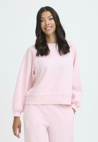 Sudadera rosa claro con mangas largas abullonadas y puños acanalados. Tela suave con un corte relajado y un dobladillo ligeramente recortado. Combinada con pantalones a juego.