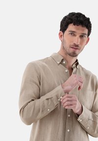 Hellbeige Hemd mit Knöpfen aus einem strukturierten Stoff, ausgestattet mit einem spitzen Kragen, langen Ärmeln und einer Brusttasche mit weißen Knöpfen.