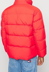 Rode pufferjack met een quilted ontwerp en hoge kraag. Heeft een gladde textuur en een losse pasvorm, gezien van achter met denim broek.