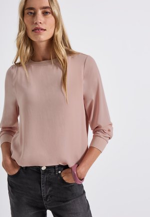 Ljusrosa långärmad blus med rundad halsringning, mjuk textur och ribbade ärmslut, matchad med mörka jeans och ett rosa armband.