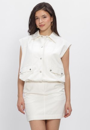 Jonge vrouw met donker haar die een witte mouwloze gilet met knopen en een witte rok draagt, staand tegen een effen lichte achtergrond.