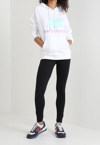 Persona che indossa una felpa bianca New Balance con logo pastello, leggings neri e sneakers navy New Balance, in piedi con le mani nelle tasche.