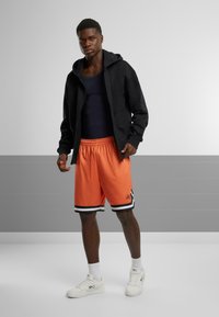 Sort zip-up hoodie, navy tanktop, orange mesh basketball shorts med sort og hvide striber, og hvide sneakers. Enkel atletisk design.