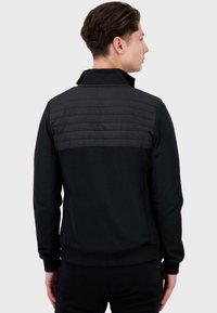 Giacca atletica nera con un pannello posteriore trapuntato testurizzato, colletto alto e polsini a coste, che offre un design sportivo e moderno.