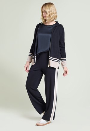 Kvinna i svart och beige randig zip-up jacka, svarta byxor med beige sidoränder och vita sneakers, står mot ljus bakgrund.