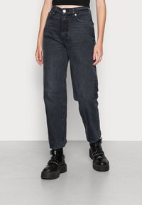 Vaqueros de mezclilla negros de talle alto con un corte relajado, cinco bolsillos y herrajes en tono cobre. Combinados con botas de plataforma negras gruesas.