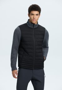 Chaleco acolchado negro con secciones aislantes, mangas acanaladas grises, cuello alto y bolsillos laterales con cremallera, llevado sobre pantalones oscuros.