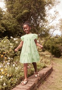 Vihreä, kerroksittain oleva mekko, jossa on röyhelöhihoja ja kukkakirjontaa. Yhdistetty kultaisiin sandaaleihin, kävelemässä tiilitalvitietä, jota ympäröivät kukat.