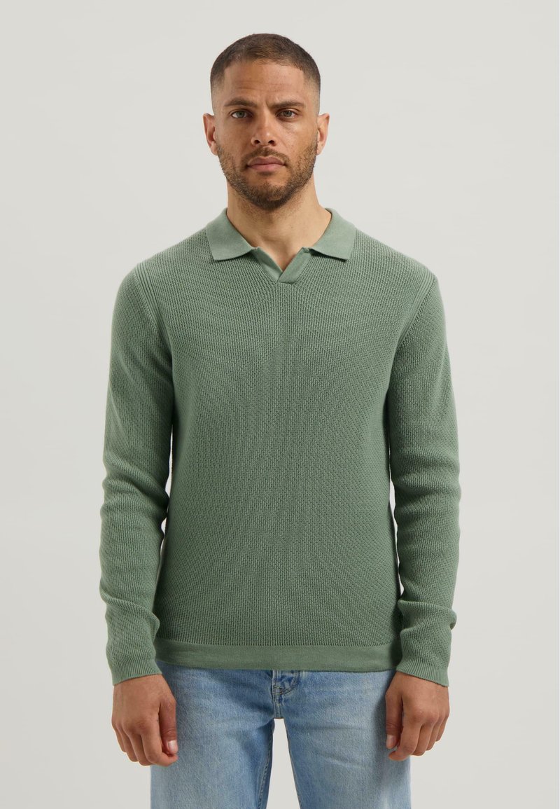 Man wearing a light green textured long-sleeve polo sweater and light blue jeans, standing against a plain light background.