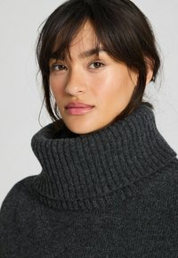 Noor naine tumedate juustega kannab paksu süsiheleda halli soonikkoega kõrge kaelusega kampsunit, vaadates veidi küljele tühja tausta vastu.