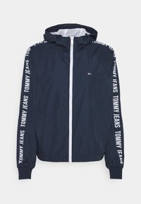 Navy blue hooded jacket with white front zipper, elastic cuffs, and "Tommy Jeans" text printed along both sleeves.