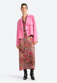 Chaqueta de mezcla de lana rosa con un diseño cropped y bolsillos frontales, superpuesta a un vestido maxi con estampado floral en tonos rosa, púrpura y marrón.