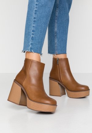 Brune læder ankelstøvler med plateau og stablede blokhæle, båret med blå cropped jeans med frynset kant på hvidt gulv.