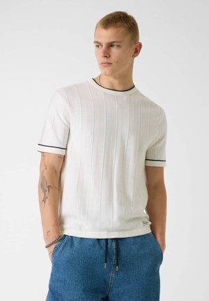 Junger Mann, der ein weißes kurzärmeliges Texturhemd mit schwarzem Rand, blaue Jeansshorts und Tätowierungen am linken Arm trägt.