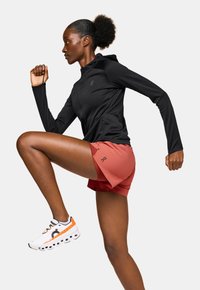 Schwarze, lange sportliche Jacke, rote Shorts und weiße Sneaker mit orangefarbenen Akzenten. Die Person befindet sich in einer Laufpose.