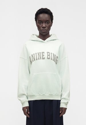 Kvinde med kort krøllet hår iført en lysegrøn hættetrøje med teksten "ANINE BING" og mørke plisserede bukser, stående mod en ensfarvet hvid baggrund.