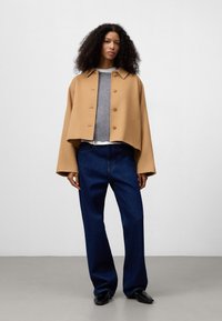Korte, camelkleurige wollen jas met een kraag en knoopsluiting, gecombineerd met een grijze trui en donkerblauwe wijde jeans.