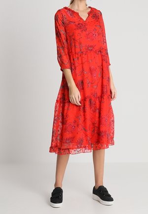 Frau trägt ein rotes Midikleid mit Blumenmuster, Dreiviertelärmeln und schwarzen Sneakers, steht vor einem schlichten hellen Hintergrund.