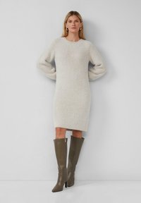 Frau steht gegen eine graue Wand und trägt ein langärmliges, cremefarbenes Strickkleid und hohe, taubfarbene Lederstiefel.