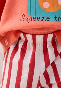 Lähivõte punaste ja valgete vertikaalsete triipudega pükstest, millel on elastne vöökoht, kantud korallikarva särgi all, millel on osaline graafika ja tekst "Squeeze."