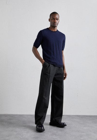 Pulover scurt din bumbac de culoare bleumarin, cu un model de wave, asortat cu pantaloni negri cu croi larg și pantofi lucioși negri, pe un fundal neutru.
