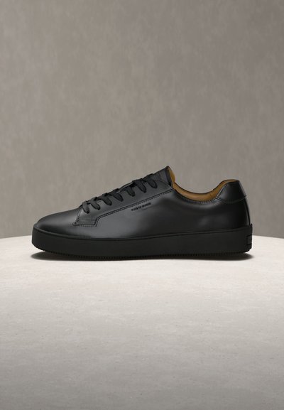 Μαύρο δερμάτινο χαμηλό sneaker με μαύρα κορδόνια, παχιά σόλα και ταμπά εσωτερική επένδυση, τοποθετημένο σε φωτεινή πέτρινη επιφάνεια.