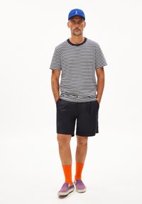 Camicia a righe a maniche corte blu scuro e bianca, abbinata a pantaloni corti blu scuro, calzini arancioni e scarpe slip-on viola. Cappellino blu in testa.