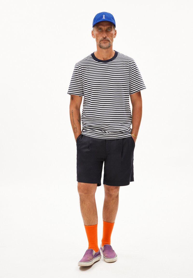 Camicia a righe a maniche corte blu scuro e bianca, abbinata a pantaloni corti blu scuro, calzini arancioni e scarpe slip-on viola. Cappellino blu in testa.