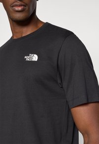 Μαύρο βαμβακερό μπλουζάκι με στρογγυλή λαιμόκοψη και κοντά μανίκια, με λευκό λογότυπο "The North Face" στο πάνω αριστε ρό μέρος του στήθους.