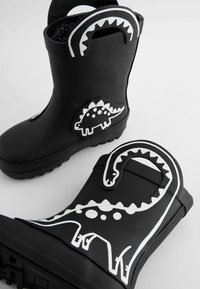Des bottes en caoutchouc noires avec des motifs de dinosaures blancs, y compris un stégosaure et une créature serpentiforme, avec des semelles texturées et des poignées latérales.
