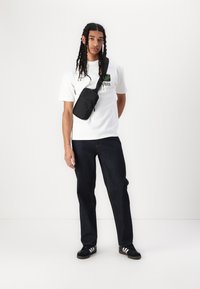 Hvit t-skjorte med grønn grafikk, svart crossover-veske, mørk denim-jeans og svarte joggesko med hvite striper, satt mot en ensfarget bakgrunn.