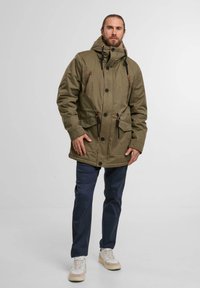 Olive green parka jacket with a hood, adjustable drawstrings, large front pockets, and button closures. Worn over dark pants and sneakers.