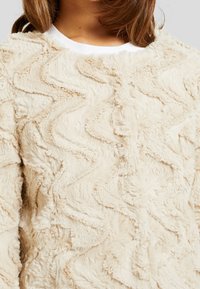 Kermanvärinen pehmeä neulepaita, jossa aaltoileva tekstuurikuvio ja pyöreä pääntie. Valmistettu pehmeästä, pörröisestä materiaalista, sopii kylmään säähän.