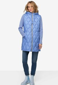 Gilet matelassé bleu clair avec capuche, doté d'une fermeture éclair intégrale, de poches latérales et de manches longues bleues. Associé à un jean bleu et des bottes de pluie.