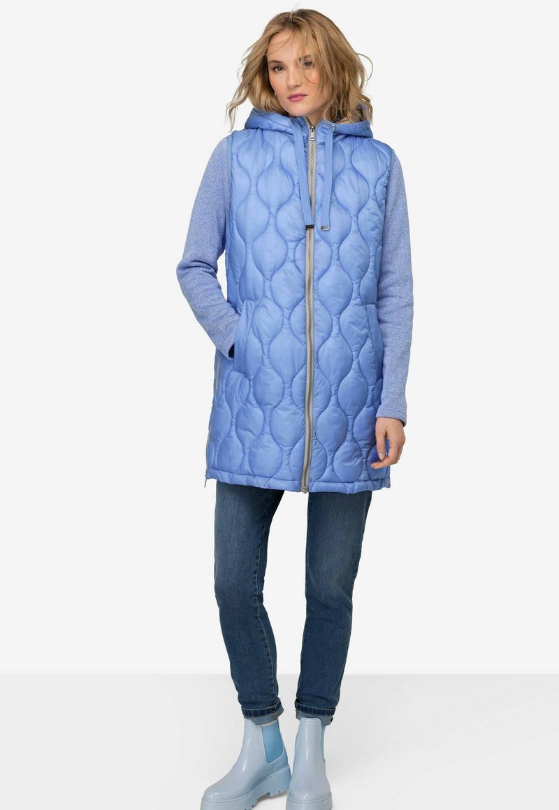 Gilet matelassé bleu clair avec capuche, doté d'une fermeture éclair intégrale, de poches latérales et de manches longues bleues. Associé à un jean bleu et des bottes de pluie.