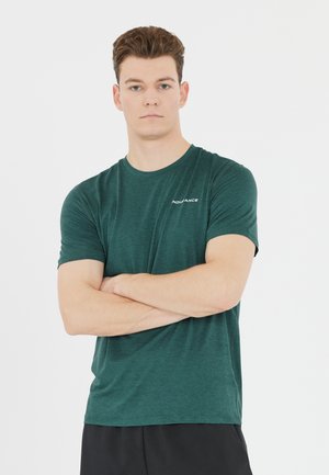 Jeune homme aux cheveux bruns et courts debout, les bras croisés, vêtu d’un t-shirt vert et d’un short noir sur fond blanc uni.