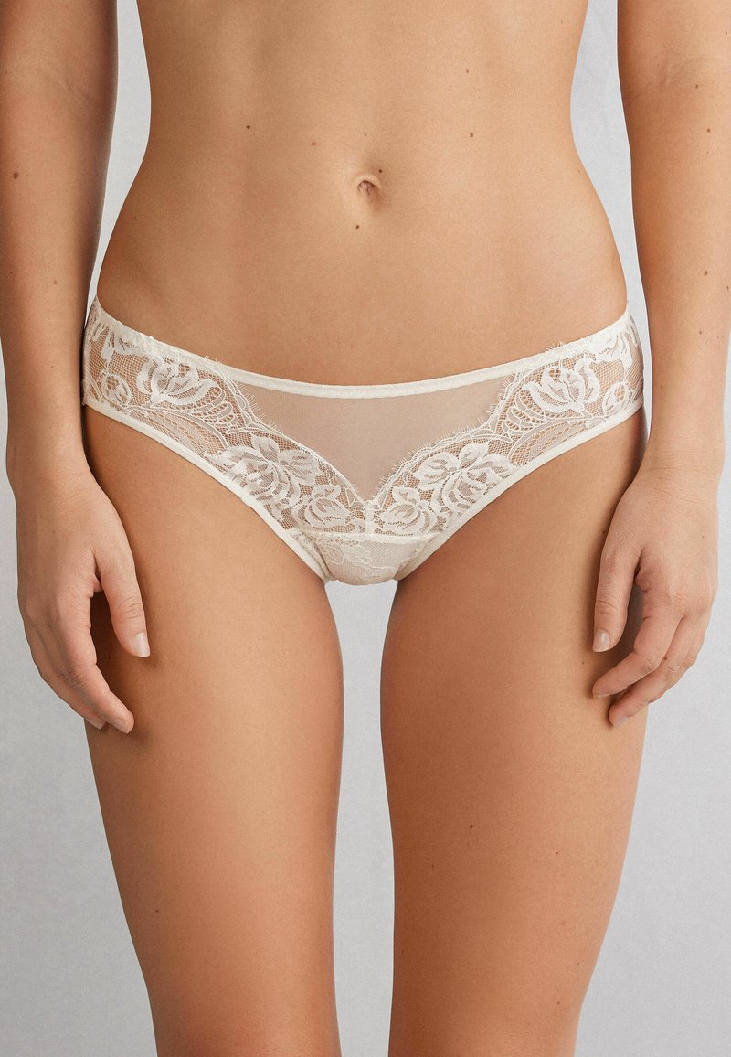 Kant bikini slipjes in licht beige, met bloemenpatronen aan de zijkanten, doorschijnende stof en een zachte elastische tailleband.