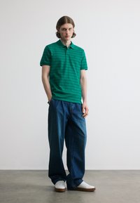 Jeune homme debout contre un mur uni, portant un polo vert à rayures, un jean bleu ample et des baskets blanches avec des semelles marron.