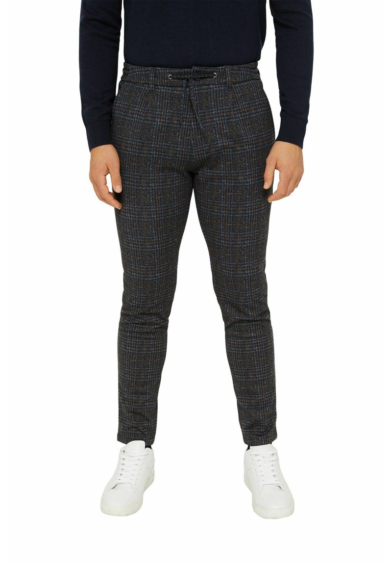 Homme portant un pantalon slim à carreaux gris foncé avec taille à cordon et des baskets blanches, debout devant un fond uni.