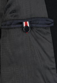 Tessuto nero con un motivo testurizzato. Presenta una chiusura a bottone e un dettaglio a nastro a strisce in rosso, bianco e blu vicino all'apertura di una tasca.