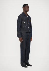 Chaqueta de mezclilla y pantalones a juego en azul oscuro, con dos bolsillos frontales, cierre de botones y detalles de costura sutiles.