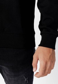 Černá žebrovaná mikina s dlouhými rukávy, s texturovaným lemem, kombinovaná s tmavými džínami z denimu s efektem opotřebení u kapsy.