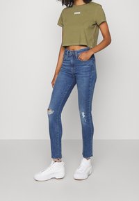 Oliivinvihreä lyhythihainen paita, jossa valkoinen logo, yhdistettynä istuviin sinisiin farkkuihin, joissa on kulutustietoja. Yhdistetty valkoisiin korkeisiin tennareihin.