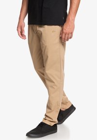 Beige chinos med en avslappnad passform, med sidofickor och en subtil logotypdetalj i ljusbrunt. Bärs med en svart skjorta och svarta skor.